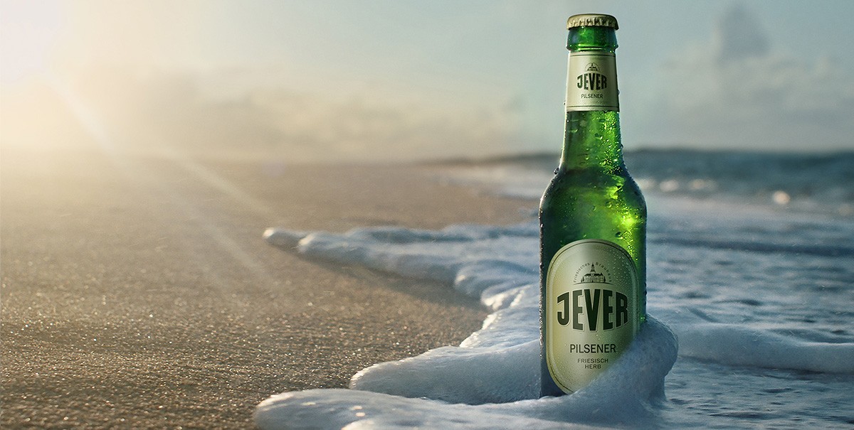 A condensation-covered beer bottle stands in the foreground on a frothy shore with a lighthouse in the background.