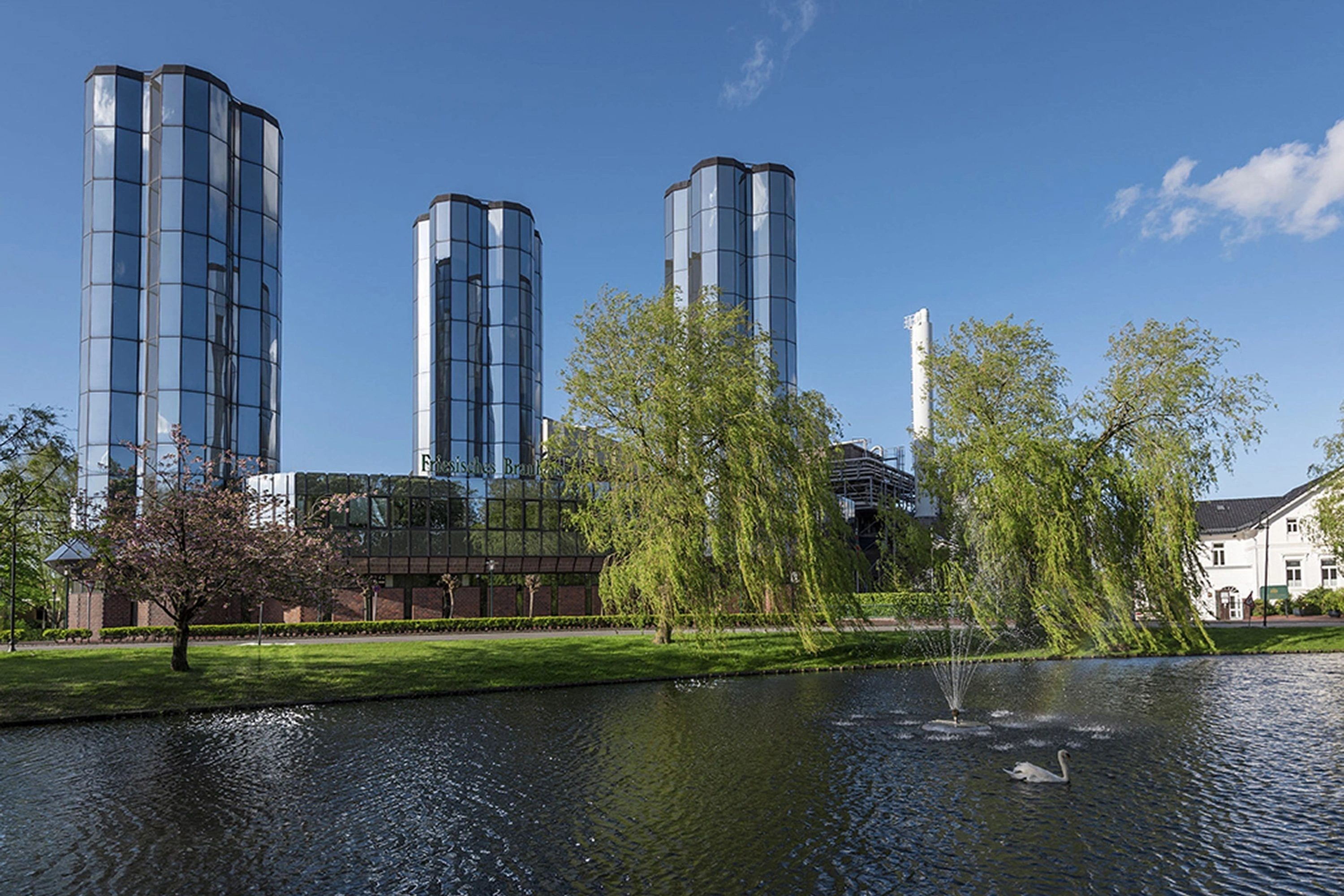Three cylindrical glass towers reflect sunlight by a tranquil pond with a fountain and a swan. Green trees frame the scene under a clear blue sky.