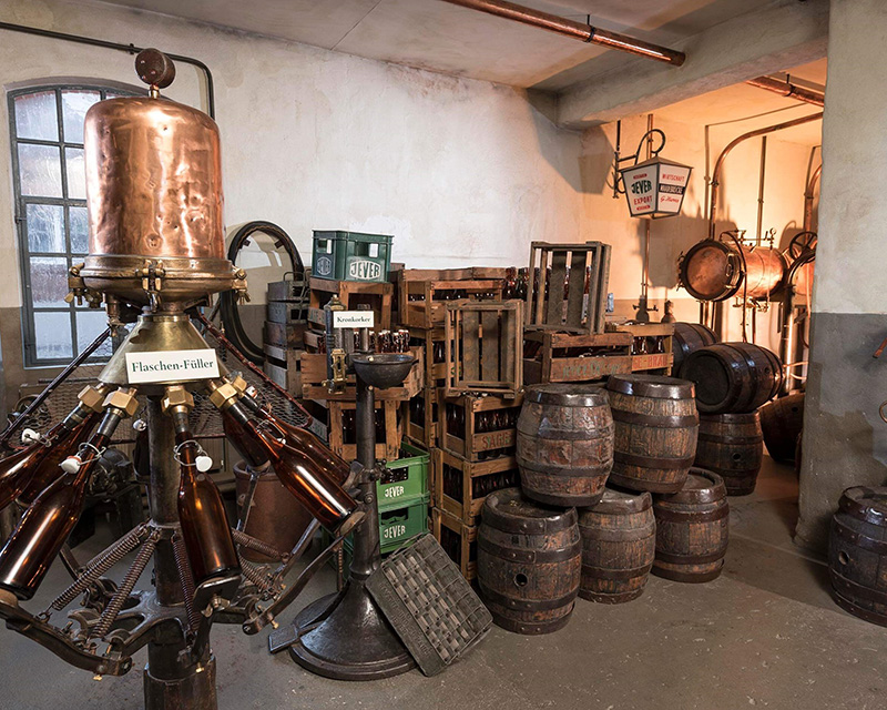 A copper distillation apparatus stands surrounded by wooden barrels and crates in a vintage brewery.