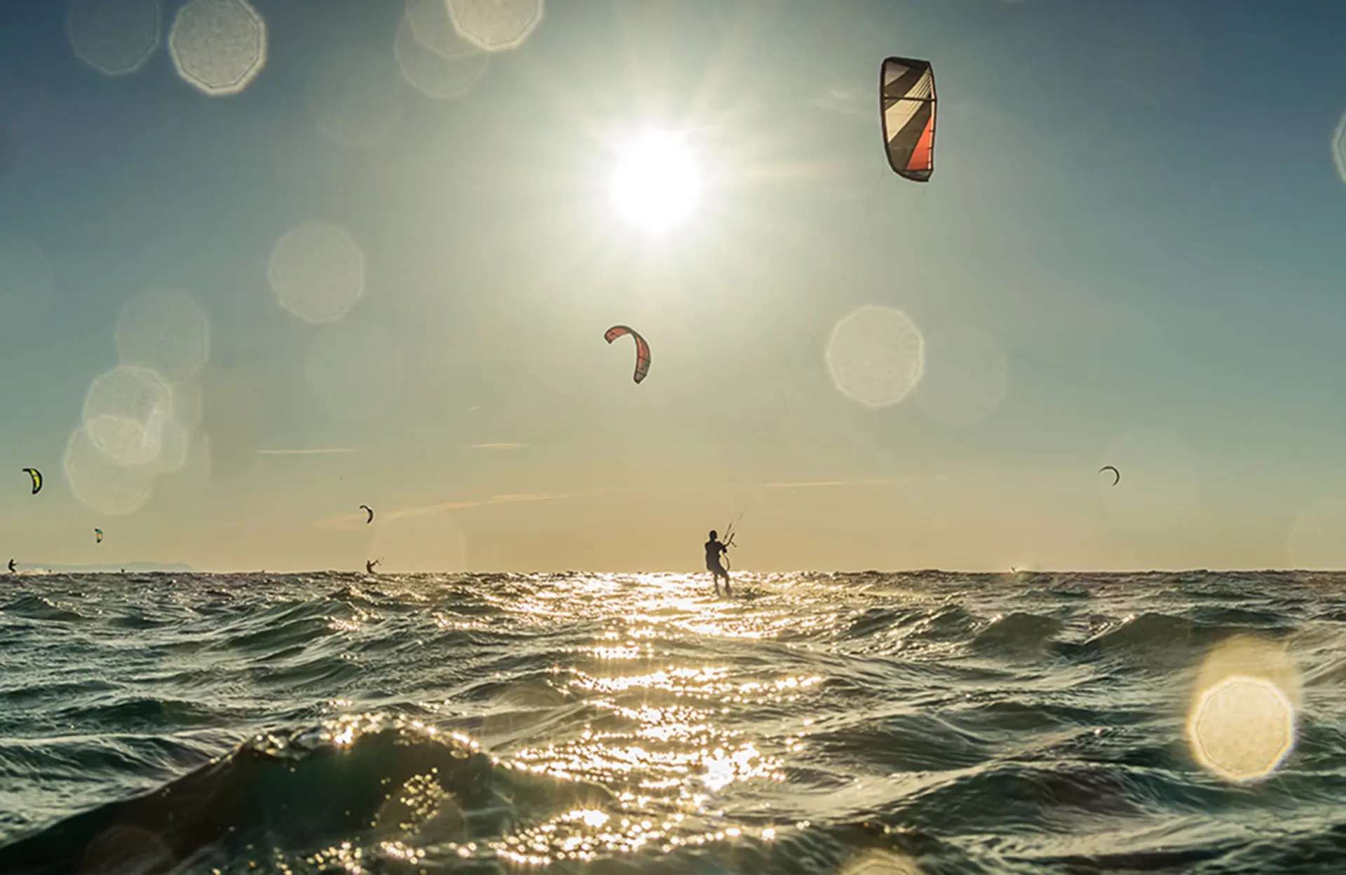 Several kite surfers glide over the ocean under a sunny sky, with light glistening on the water.