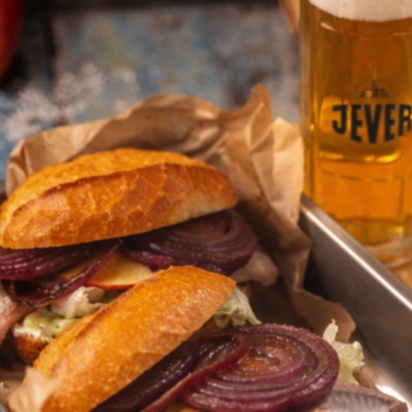 A sandwich with red onions, lettuce, and sliced meat is served next to a glass of beer.