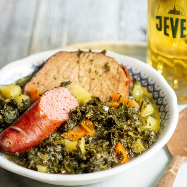 A bowl of kale stew with slices of sausage and a side of bread, served with a glass of beer.