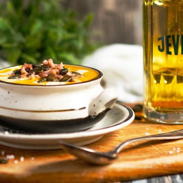 A bowl of creamy soup is topped with seeds and bacon bits, served next to a glass of beer on a wooden table.