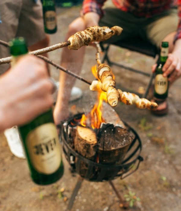Two people are roasting dough on sticks over a campfire, with bottles of beer nearby.