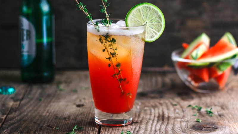 A tall glass of iced red cocktail garnished with lime and thyme sits on a wooden table.