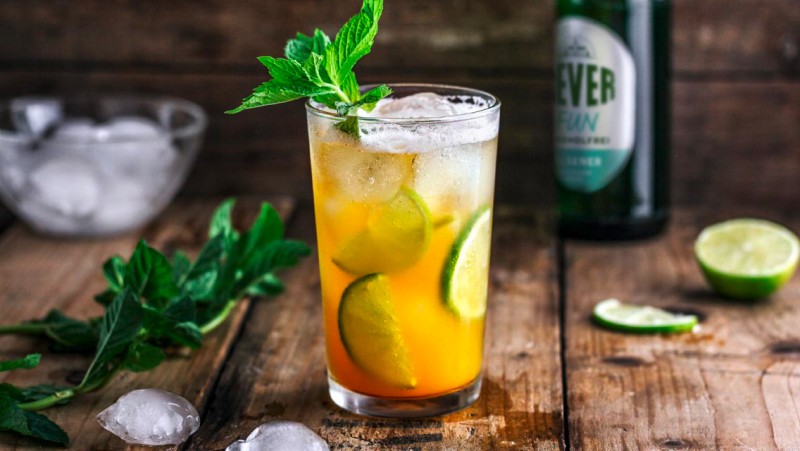 A glass filled with ice and an orange-colored liquid is sitting on a wooden table. It is garnished with lime slices and mint.