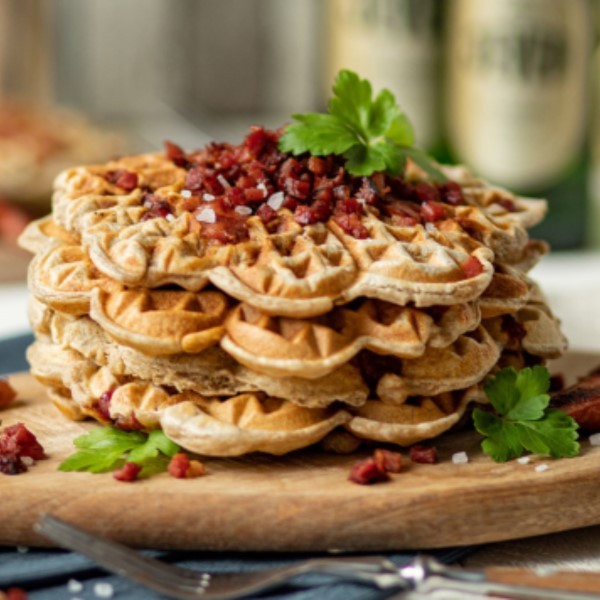 A stack of waffles is topped with diced bacon and garnished with parsley on a wooden board.