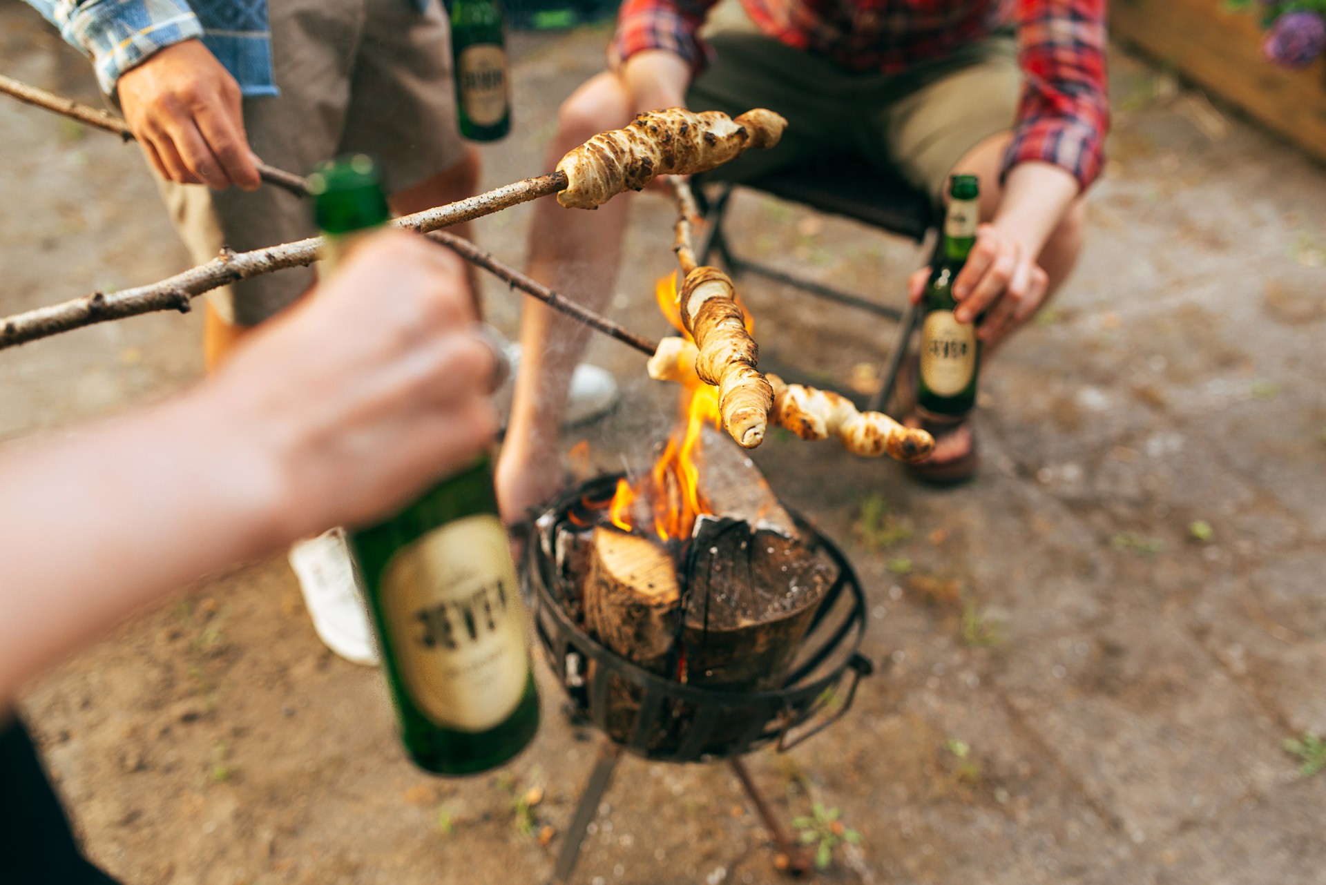 Three people are toasting with bottles while roasting dough on sticks over a campfire.