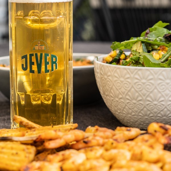 A glass of beer stands next to a bowl of salad and grilled shrimp on a table.
